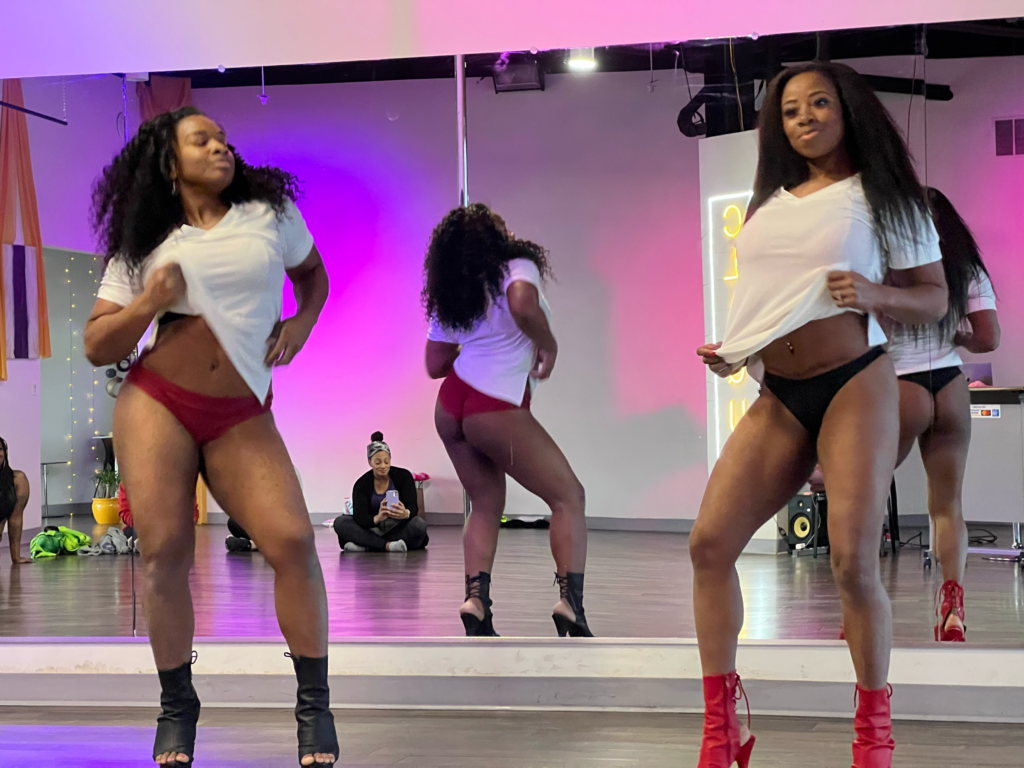 Two African American women dressed in what shirts with black and red heels and pole shorts and heels as they practice for their performance during Slay the Stage rehearsal 